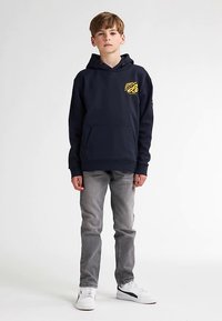 Námořnicky modrá mikina s kapucí a přední kapsou, na hrudi žlutý grafický motiv, doplněná šedými džínami a bílými teniskami. Hladká textura látky.