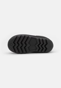 Semelle noire d'une chaussure présentant des motifs de crampons en zigzag profonds et une zone hexagonale avec le logo « UGG » embossé au centre.