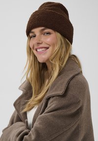 Căciulă texturată maro cu un vârf rotunjit, asortată cu un palton gri, fără guler. Persoana care o poartă are păr lung, de culoare castanie deschisă.