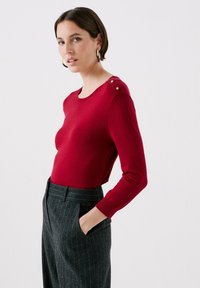 Röd långärmad topp med rund halsringning och knappryggsdetaljer på axeln, i kombination med grå ränderbyxor. Slät textur.