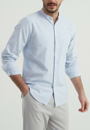 Chemise boutonnée rayée bleu clair avec col montant, manches longues retroussées et texture légère, associée à un pantalon gris clair.