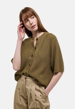 Femme aux cheveux bruns lisses portant un cardigan texturé vert olive à boutons et un pantalon beige plissé, posant avec une main dans la poche.