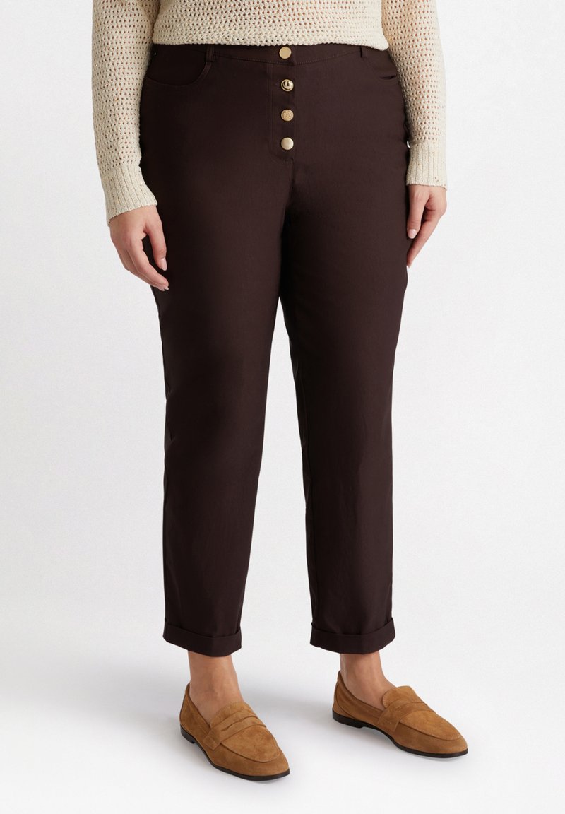 Pantaloni marroni in cotone con un design aderente, dotati di una chiusura a cinque bottoni, passanti per la cintura e orli arrotolati, abbinati a mocassini beige.