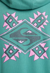 Green hoodie featuring a large geometric graphic in pink and gray. The design includes spikes and diamond shapes, with textured detailing.