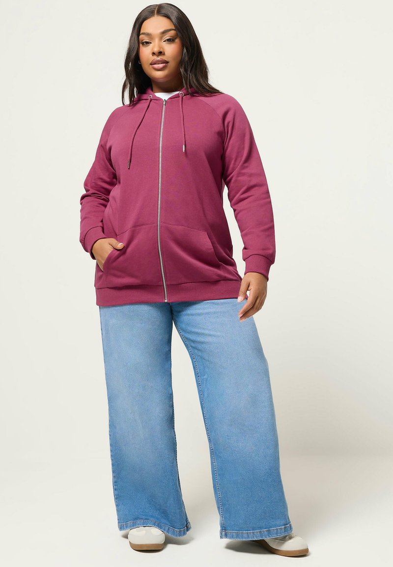 Donna in piedi che indossa una felpa con cappuccio marrone con zip, jeans a gamba larga di colore azzurro chiaro e sneakers bianche, su uno sfondo chiaro e semplice.