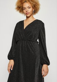 Μαύρο φόρεμα με μακριά μανίκια από υφαντό γυαλιστερό ύφασμα, με V λαιμόκοψη, συγκεντρωμένη μέση και ογκώδη μανίκια. Ασημί σκουλαρίκια προσθέτουν λεπτομέρεια.