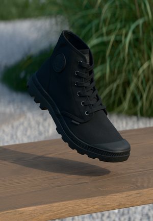 Sort højtop sneaker med tyk sål og snørebånd, svævende over en træoverflade, med grønne planter og grus i baggrunden.