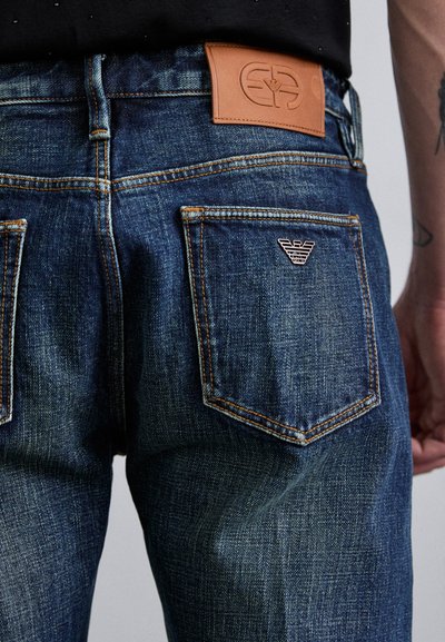 Tamsiai mėlyni džinsai su odiniu užsegimu juosmens srityje, oranžinėmis siūlėmis ir mažu metaliniu logotipu ant galinio kišenės.