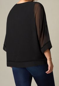 Czarna przezroczysta bluzka o luźnym kroju, z szerokimi rękawami 3/4 oraz misternym zdobieniem z koronki wzdłuż krawędzi dołu i rękawów. Noszona na niebieskich dżinsach.