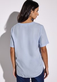 Blusa azzurro chiaro a maniche corte con scollatura arrotondata e risvolti, caratterizzata da una trama liscia e un'orlo posteriore leggermente più lungo.