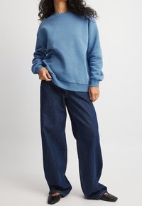Persona con pelo rizado que lleva un sudadera azul, jeans oscuros de pierna ancha y zapatos de tacón puntiagudos negros, de pie contra un fondo sencillo.