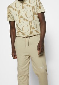 Beige bomulls t-shirt med ett stort CK-logomönster, kombinerad med matchande beige sweatpants som har en liten vit "Calvin Klein Jeans"-logotyp.