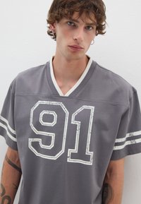 Jersey gris con detalles en blanco, con el número "91" en impresión desgastada. Mangas cortas, cuello en V, tejido de mezcla de algodón.