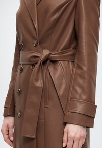 Καφέ δερμάτινο παλτό λάβας με διπλή σειρά κουμπιών και δεμένη ζώνη, φορεμένο από άτομο με εμφανή χέρια στα πλάγια.