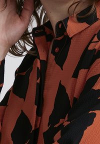 Camicetta a maniche lunghe in tessuto rosso-arancione texture con motivi astratti neri. Presenta un colletto e una fila di bottoni.