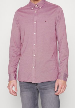 Mand iført en langærmet knappet mauve skjorte med et lille Tommy Hilfiger-logo på brystet og grå jeans.