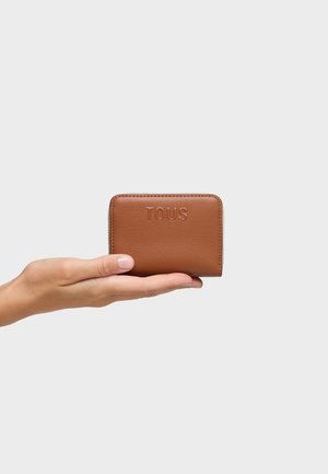 Mano che tiene un piccolo portafoglio marrone in pelle con logo "TOUS" in rilievo e chiusura con zip su uno sfondo bianco semplice.