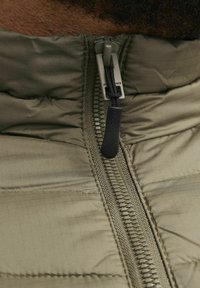 Olijfgroene pufferjack met ritssluiting. Getextureerd materiaal, lineair patroon zichtbaar, stevige rits met een stoffen trekband.
