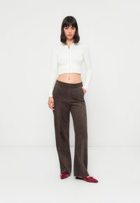 Frau mit dunklen Haaren, die einen weißen, kurzen Reißverschluss-Pullover, braune weite Hosen und rote Ballerinas trägt, steht vor einem schlichten weißen Hintergrund.