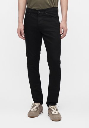 Mann trägt schwarze Slim-Fit-Jeans, graue Wildleder-Sneakers und ein olivgrünes Hemd, steht vor weißem Hintergrund.
