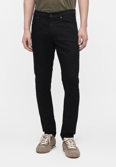 Mand iført sort slim-fit jeans med grå ruskindssneakers og en olivengrøn skjorte, står mod en hvid baggrund.