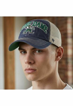 Young man wearing a navy and beige mesh Notre Dame baseball cap, looking forward with a neutral expression indoors.