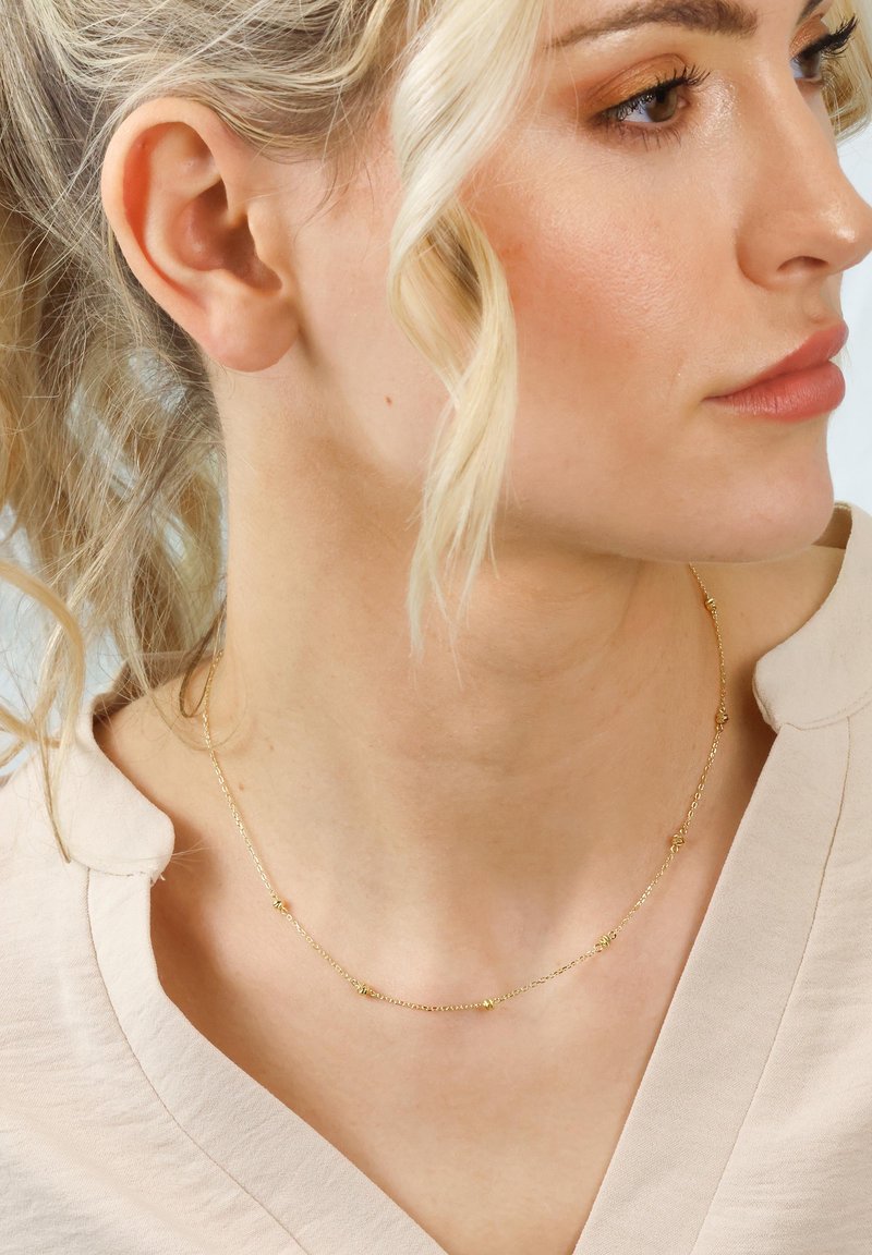 Nahaufnahme eines Frauenhalses und -gesichts, das eine zarte goldene Halskette mit kleinen Perlenakzenten und einer hellbeigen Bluse zeigt.