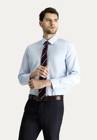 Lichtblauw overhemd met een puntkraag, gecombineerd met een donker gestreepte stropdas en donkere broek. Voorzien van knoopmanchetten en een bruine riem.