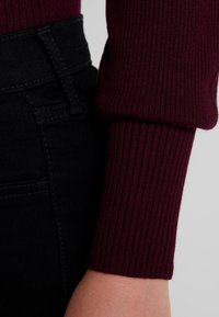 Burgunderfarbener, gerippter Pulloverärmeln, kombiniert mit schwarzen hochtaillierten Jeans, die eine glatte Textur und dezente Nähdetails aufweisen.