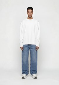 Νέος άνδρας που στέκεται προς τα εμπρός, φορώντας λευκό πουκάμισο με μακριά μανίκια, χαλαρά μπλε τζιν και λευκά αθλητικά παπούτσια, μπροστά σε έναν απλό ανοιχτόχρωμο φόντο.