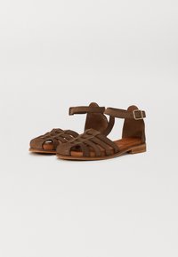 Sandales plates en suède marron avec un design de lanières entrecroisées, une boucle à la cheville et une semelle en bois plate. Elles possèdent une semelle intérieure texturée pour une meilleure adhérence.