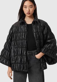 Svart puffjacka med en glansig textur, detaljerad med samlad sömnad, hög krage och dragkedja fram. Kombinerad med en svart topp och mörka jeans.