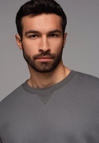 Sudadera gris con cuello redondo, tela texturizada y un pequeño detalle de costura en forma de triángulo en el collar, llevada por un modelo.