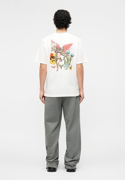 Άνδρας με σγουρά μαλλιά στέκεται πλάτη φορώντας λευκό μπλουζάκι με πολύχρωμα γραφικά αγγέλου, κάκτου και κειμένου, καθώς και χαλαρό γκρι παντελόνι.