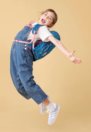 Mädchen springt mit ausgebreiteten Armen, trägt gestreifte Latzhose, ein weißes T-Shirt, blaue Turnschuhe und einen blauen gepunkteten Rucksack vor einem beigen Hintergrund.