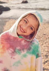 Hætteponcho i blødt stof med et tie-dye-mønster i pastelfarver: lyserød, grøn, gul og lilla. Blød tekstur, løs pasform.