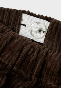 Ceinture en velours côtelé brun avec une sangle blanche réglable. Comprend une fermeture à bouton en plastique transparent. Tissu doux et texturé avec des côtes verticales.