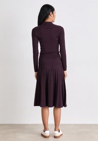 Tamno ljubičasta haljina sa rebrastim uzorkom, prilagodljivim dugim rukavima, plisiranom suknjom i bijelim tenisicama. Materijal izgleda mekano s glatkom teksturom.