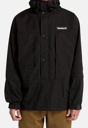 Veste à capuche noire Timberland avec pressions à l'avant et poignets élastiques portée par une personne aux mains tatouées et pantalon marron.