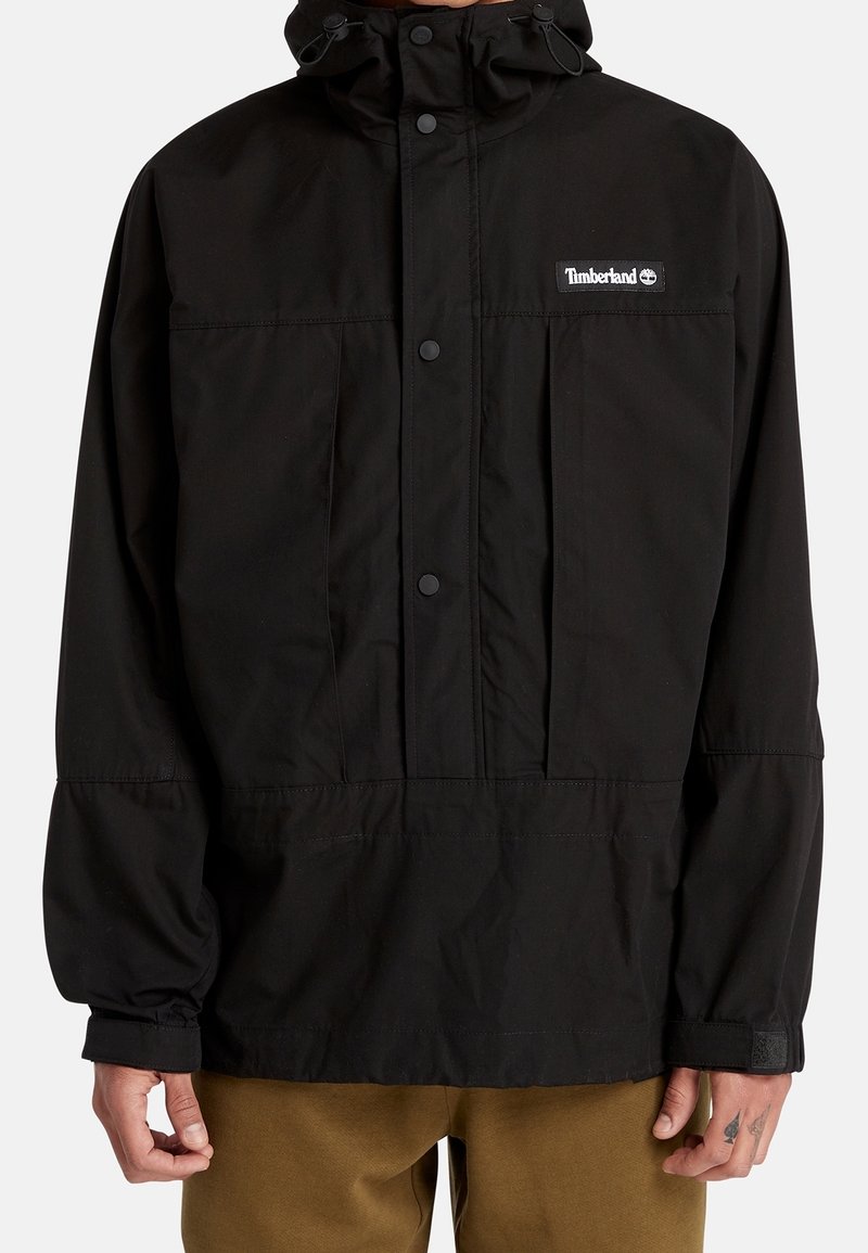 Veste à capuche noire Timberland avec pressions à l'avant et poignets élastiques portée par une personne aux mains tatouées et pantalon marron.