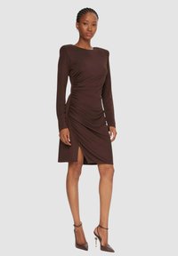Abito marrone a maniche lunghe con silhouette aderente, dettagli arricciati e spacco laterale, abbinato a scarpe marroni con tacco alto.