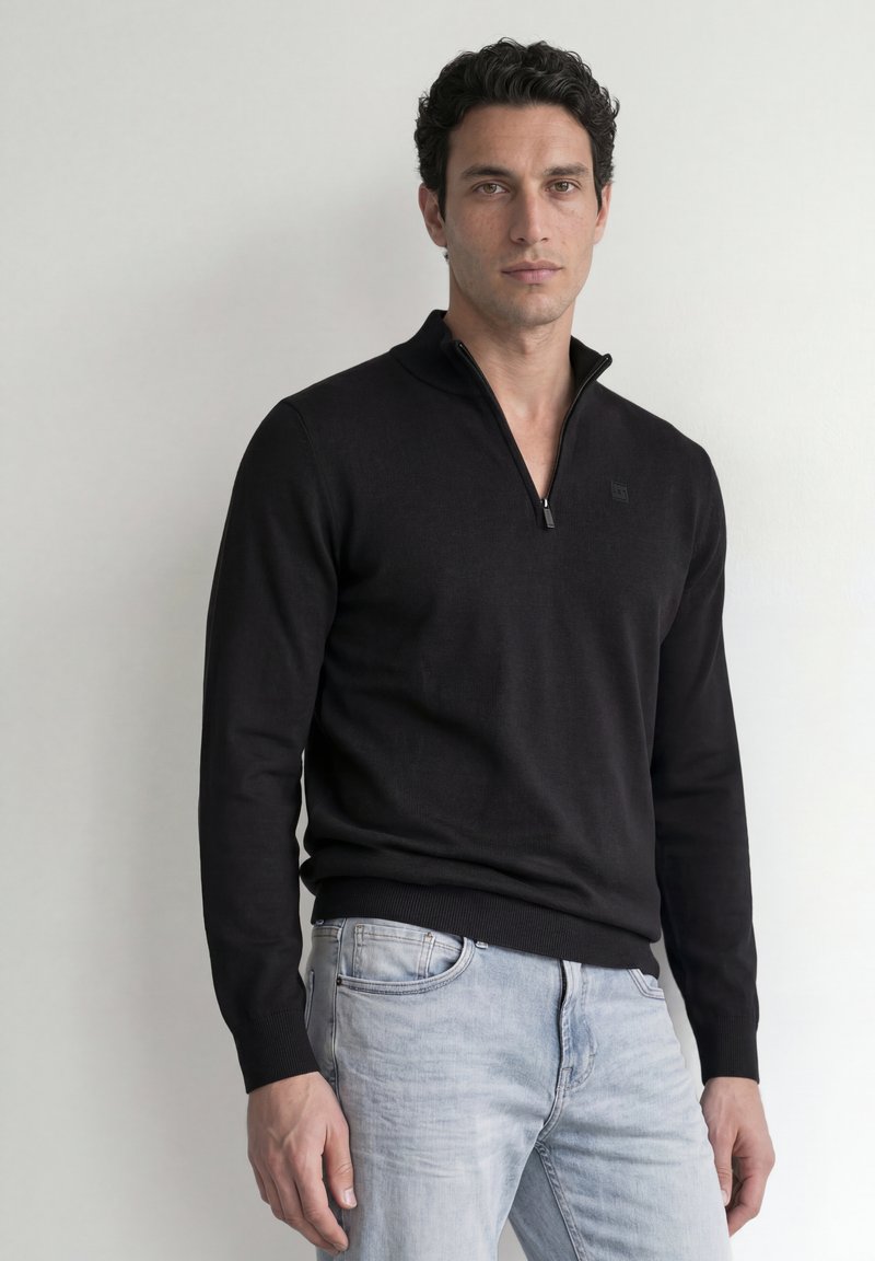 Homme aux cheveux bouclés et foncés portant un pull noir zippé et un jean bleu clair, debout contre un mur blanc.