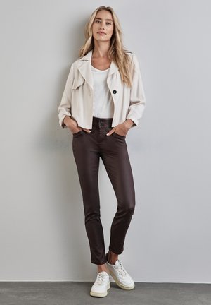 Giacca cropped beige chiaro con un singolo bottone nero, sopra una maglietta bianca, abbinata a pantaloni aderenti marroni e sneakers bianche.