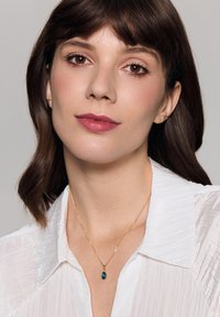 Złoty naszyjnik z łańcuszkiem i łzowym turkusowym pendanem, noszony na białej, teksturowanej bluzce, podkreślający biżuterię na tle tkaniny.