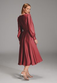 Bordeaux Satin-Kleid mit langen Puffärmeln, taillierter Taille und ausgestelltem Rock. Silberne, abgeschnallte hohe Sandalen runden den Look ab.