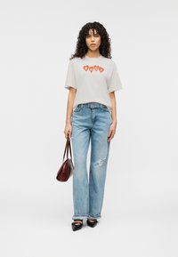 Ανοιχτό γκρι μπλουζάκι με κόκκινα γράμματα "HUGO", συνδυασμένο με ανοιχτό μπλε τζιν παντελόνι φαρδύ στο πόδι με φθαρμένη λεπτομέρεια. Αξεσουάρ περιλαμβάνουν μικρή καφέ τσάντα.