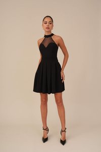 Abito nero con corpino aderente e gonna plissettata, caratterizzato da una scollatura in rete trasparente e un design allacciato al collo che mette in risalto la silhouette. Indossato con sandali con i lacci.