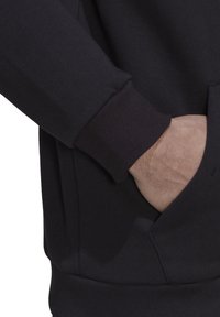 Felpa nera con tasca a marsupio, polsini ribattuti e una morbida texture. Ingrandimento di una mano poggiata nella tasca e dettaglio della manica.