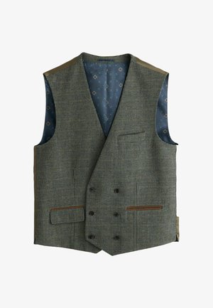 Mand iført grå vest og bukser med hvid skjorte og beige slips, står foran en stor bronzedør med et lodret håndtag.