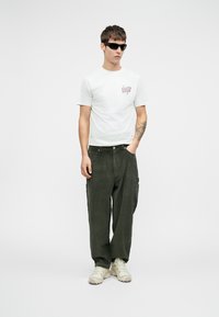 Biela bavlnená tričko s grafickým potlačou, skombinované so zelenými kordovými nohavicami. Outfit obsahuje tenisky neutrálnych farieb a slnečné okuliare.